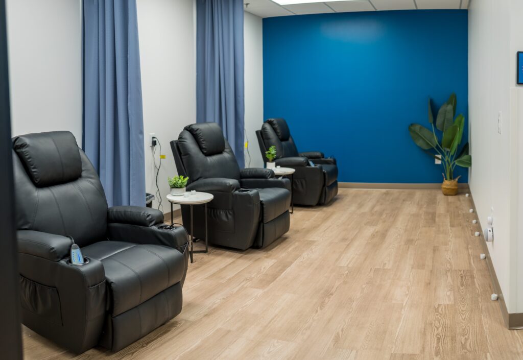 Modern patient recovery or consultation room featuring several black leather recliners, wood flooring, blue accent wall, and a minimalist decor style with potted plants.