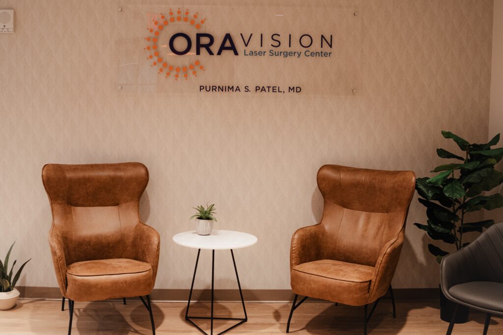 Cozy waiting area at ORA Vision with two brown armchairs, a small white round table, a potted plant, and a wall sign displaying the clinic's name and the doctor’s name