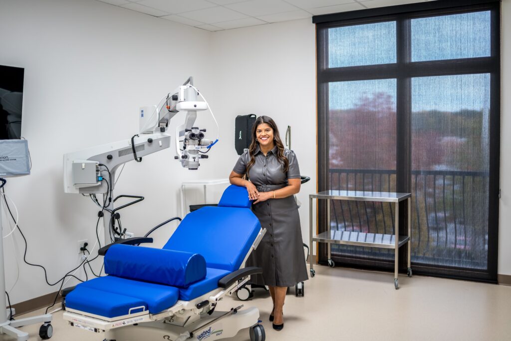 A female eye doctor standing beside a blue ophthalmic exam chair and surgical equipment in a brightly lit examination room with large windows.